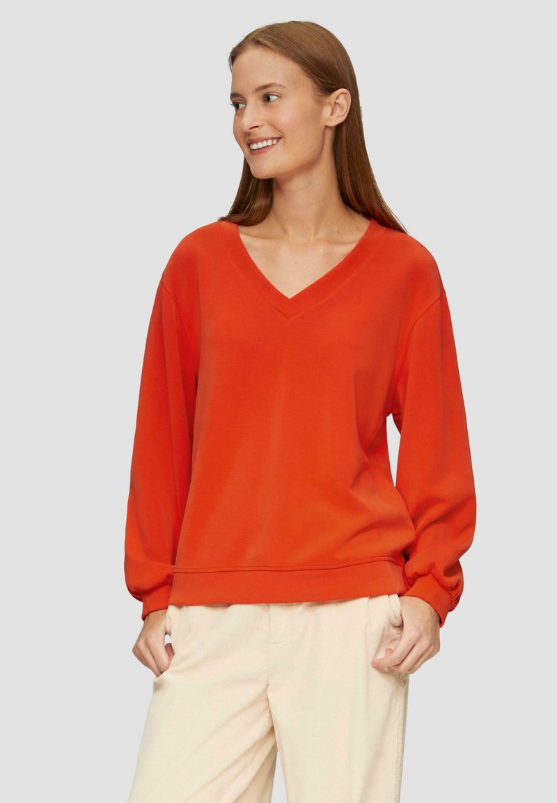 Czerwony sweatshirt z dekoltem w serek, z długimi, marszczonymi rękawami i luźnym krojem. Gładka faktura, ściągacz u dołu i przy mankietach. Noszony z jasnymi spodniami.