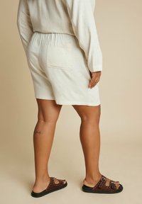 Vaaleanbeige pellavashortsit, joissa on joustava vyötärö ja takatasku. Ruskeat sandaalit, joissa on koristeellisia niittejä ja tasapohja.