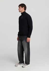 Maglione nero con zip, jeans grigi e sneakers di colore chiaro. Il maglione ha polsini e orlo a coste; i jeans hanno un fitting rilassato con una texture sbiadita.