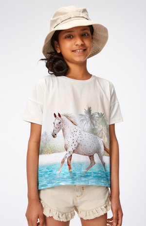 Camicetta a maniche corte di colore crema con una grafica di un cavallo bianco che saltella su uno sfondo di spiaggia. Abbinata a pantaloni corti sfilacciati beige.