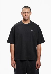 Homme avec les cheveux courts et des boucles d'oreilles portant un T-shirt noir ample avec le texte "FAVELA", se tenant devant un fond blanc uni.