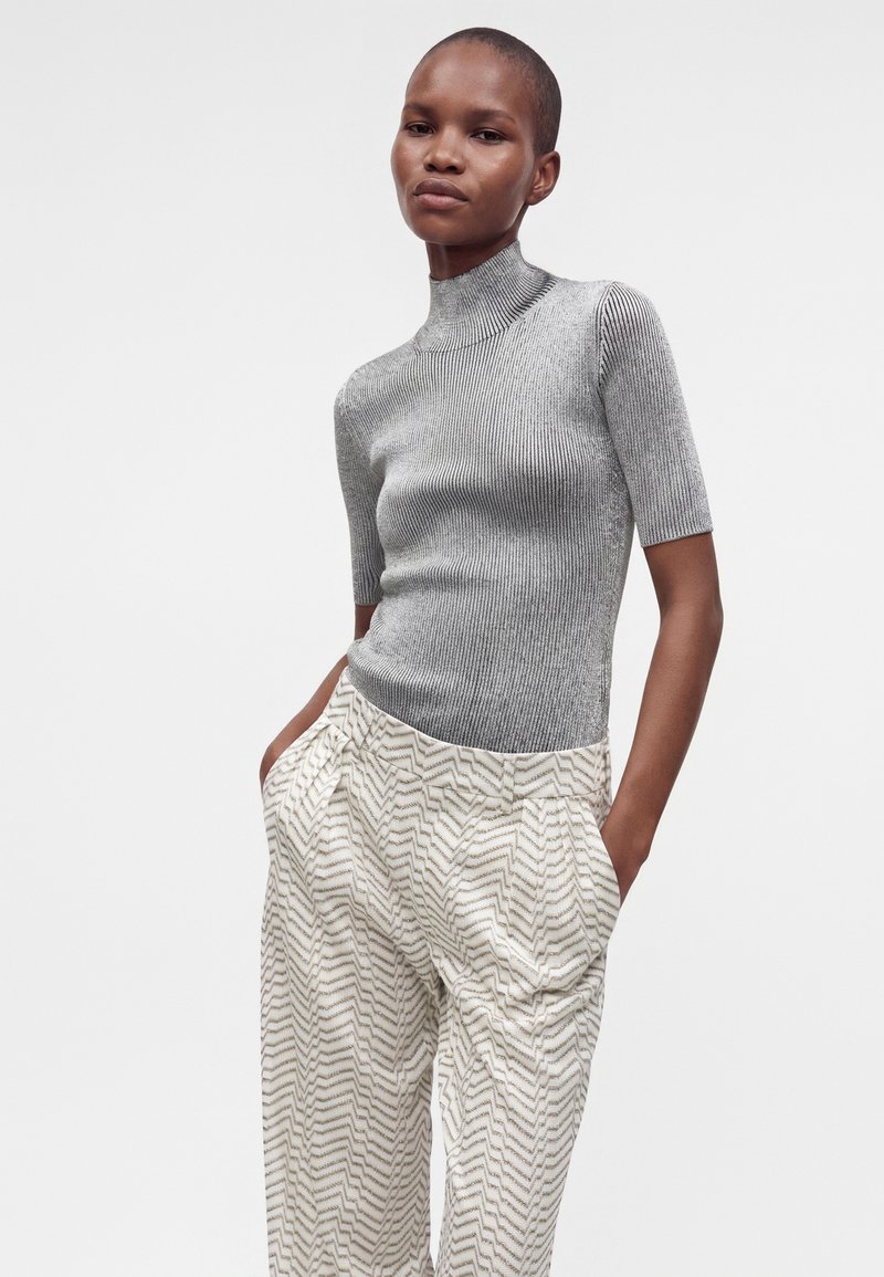Femme aux cheveux courts portant un haut gris côtelé à col roulé ajusté et un pantalon blanc ample avec un motif en zigzag, les mains dans les poches.