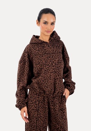 Kobieta w brązowo-czarnym kapturze w panterkę i dopasowanych spodniach stojąca na białym tle.