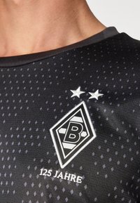 Svart träningsskjorta med ett subtilt prickmönster, försedd med en vit diamantformad logotyp med ett 'B', två stjärnor ovanför och texten "125 Jahre" nedanför.