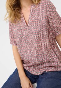 Krótki rękaw bluzki wykonanej z lekkiego materiału w różowo-granatowy wzór w zygzak, z dekoltem w kształcie litery V i luźnym krojem.
