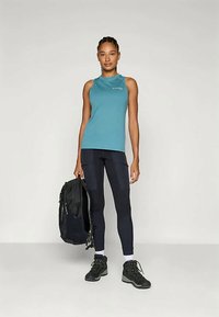 Mujer joven de pie sosteniendo una mochila negra, vistiendo una blusa sin mangas color teal, pantalones cargo negros, calcetines blancos y botas de senderismo negras.