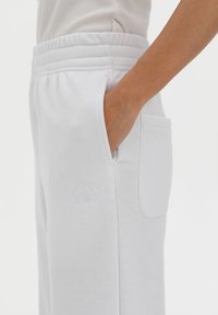 White sweatpants with an elastic waistband, side pocket, and a subtle logo embroidered on the lower leg. Soft fabric with a smooth texture.