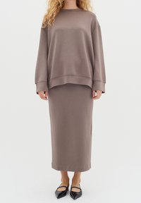 Sudadera marrón de punto con mangas largas y cuello redondo combinada con una falda larga a juego. Zapatos planos negros de punta puntiaguda.