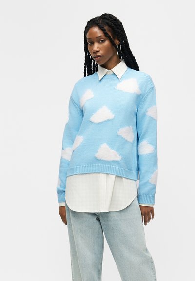 Femme aux longs cheveux tressés portant un pull bleu clair avec des motifs de nuages blancs par-dessus une chemise blanche à col, et un jean bleu clair, debout devant un fond uni.
