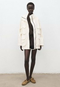 Witte oversized jas met borst- en zijzakken, gecombineerd met een donkerbruin topje en shorts, en lichtbruine veterschoenen. Effen achtergrond.
