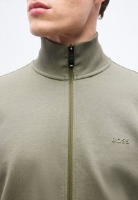 Άνδρας με ανοιχτό λαδί ζακέτα με φερμουάρ, ριμπ ψηλό γιακά και το λογότυπο "BOSS" στο στήθος, κοιτάζοντας προς τα αριστερά, περικομμένος κάτω από τη μύτη.