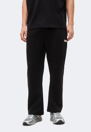 Άτομο που φορά μαύρο παντελόνι άνετης εφαρμογής με μικρό λογότυπο στον μηρό και αθλητικά ασημί-γκρι παπούτσια, στέκεται μπροστά σε απλό φόντο.