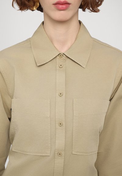 Camisa beige de botones con cuello, que presenta dos bolsillos de parche en el frente y una textura sutil. Diseño clásico con detalles simples y funcionales.