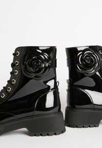 Botas de tobillo de charol negro con suela gruesa, que presentan acentos decorativos de rosas negras en los laterales y ojales de hardware dorado.