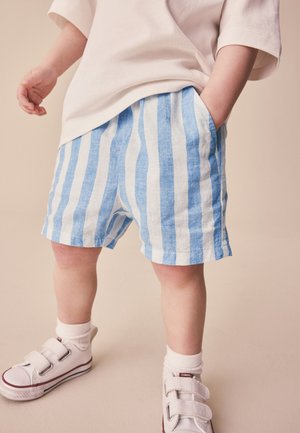 Kind trägt blau-weiß gestreifte Shorts, beige Hemd, weiße Socken und weiße Klettverschluss-Sneakers und steht auf beigem Hintergrund.