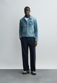 Muž v modrej rifľovej bunde s potlačou, bielom tričku, tmavých širokých nohaviciach a krémových topánkach stojaci pri jednofarebnej svetlej stene.