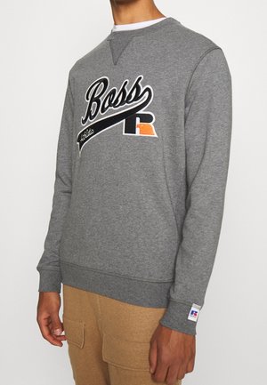 Mand iført en grå sweatshirt med "Boss Athletic" logo og beige cargobukser, stående foran en ensfarvet hvid baggrund.