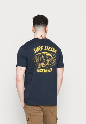 Mężczyzna z krótkimi kręconymi włosami i bielactwem, ubrany w granatowy T-shirt z żółtą grafiką surfingową i napisem "Surf Siesta Quiksilver" na plecach.