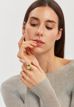 Frau mit langen braunen Haaren, die einen silbernen Pullover trägt, berührt ihr Gesicht und zeigt einen großen, glänzenden goldenen Ring an ihrem Finger.