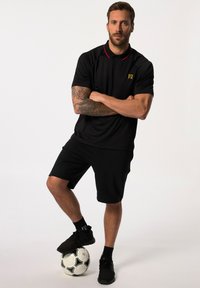 Polo atletico nero con accenti rossi, abbinato a pantaloni corti neri. Presenta un logo sul lato sinistro del petto. Il modello è in piedi su una palla da calcio.