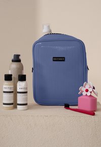 Blauwe harde reiskoffer met ribbeltextuur, zwart logo, omgeven door flessen shampoo en conditioner, een roze opbergdoos en een rode tandenborstel.