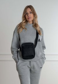 Sort crossbody taske med struktureret stof, rektangulær form og justerbar rem, båret over en grå hættetrøje og sweatpants.