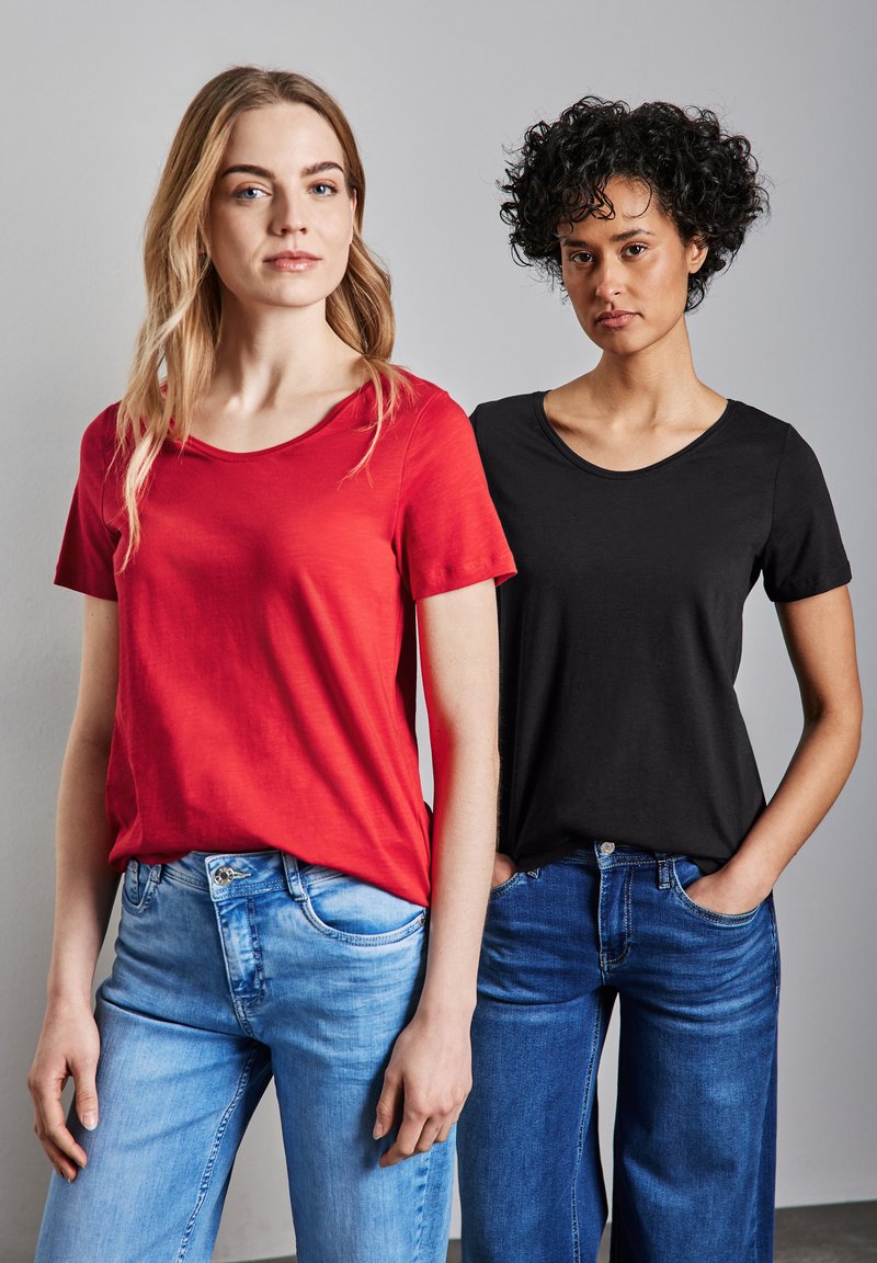 Dos mujeres están de pie una al lado de la otra, vistiendo camisetas. La camiseta de la izquierda es roja, la de la derecha es negra. Ambas camisetas son de manga corta y tienen cuellos redondos.