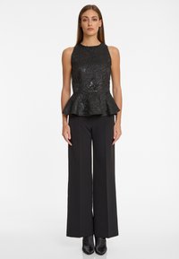 Femme portant un top péplum noir à motifs sans manches, un pantalon noir à jambe large et des bottes noires à bout pointu, se tenant contre un fond blanc.