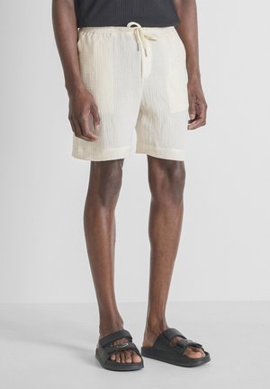 Homme portant un short texturé couleur crème avec taille à cordon et poches avant, sandales slides noires, debout sur un sol gris.