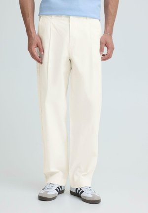 Mees, kes kannab lahtisi valgeid pükse, helehalli sinist särk ja valgeid tossusid mustade triipudega, seisab ühevärvilise hele tausta ees.