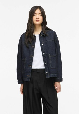 Jeune femme aux longs cheveux foncés portant une veste en jean foncée par-dessus une chemise blanche et un pantalon noir à jambes larges sur un fond blanc uni.