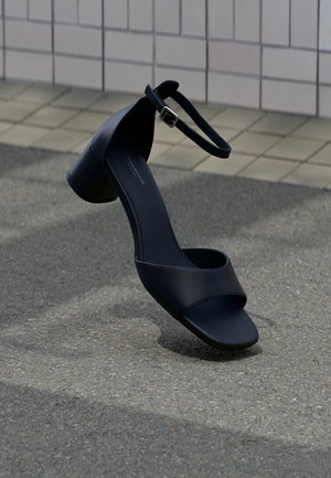 Sort læderlavhælet sandal med ankrem og spænde, placeret oprejst på en flise- og asfaltflade.