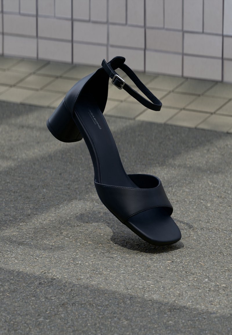 Sandale noire en cuir à petit talon avec bride de cheville et boucle, positionnée debout sur une surface carrelée et asphaltée.