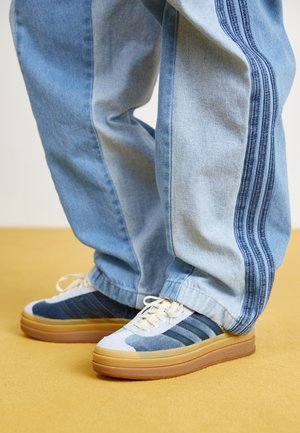Niebiesko-białe sneakersy z gumowymi podeszwami noszone z luźnymi jasnoniebieskimi jeansami z ciemnoniebieskimi potrójnymi paskami po bokach.