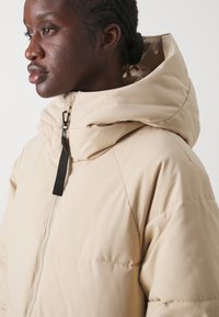 Veste matelassée beige avec un col montant, une capuche ajustable et une fermeture éclair sur le devant. Elle présente un tissu texturé et des accents matériels foncés.