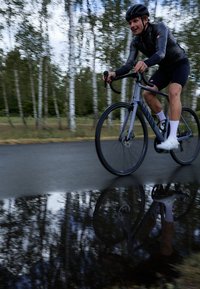 En cyklist i svart hjälm och åtsittande svart cykelutrustning cyklar på en grå cykel på en våt väg, som reflekterar träd i en vattenpöl bredvid stigen.