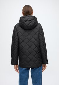 Μαύρο καπιτονέ μπουφάν με κουκούλα, με μοτίβο διαμαντιού, μακριά μανίκια και άνετη εφαρμογή, φορεμένο πάνω από μπλε τζιν.