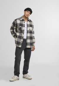 Rutete flanellskjorte i krem, svart og grå med to brystlommer; grå bukser; hvite sneakers med svarte detaljer; svart lue.