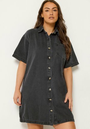 Kobieta o długich brązowych włosach, ubrana w luźną, krótką, czarną sukienkę jeansową z guzikiem i kieszenią na piersi, stojąca na jednolitym tle.