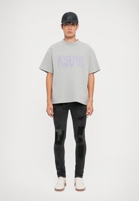 T-shirt grigia oversize con stampa viola "KSUBI". Jeans skinny neri consumati con toppe. Sneakers beige. Cappellino da baseball nero.