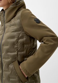 Oliwkowa kurtka z pikowanym korpusem, gładkimi rękawami i kapturem. Posiada czarną naszywkę z logo na lewym rękawie oraz boczne kieszenie.