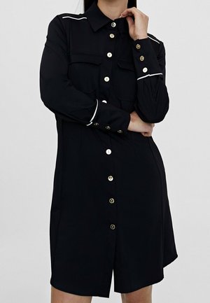 Kvinde iført en sort skjortekjole med hvide kanter på manchetter og skuldre, poserer med den ene hånd på hagen og den anden arm krydset.