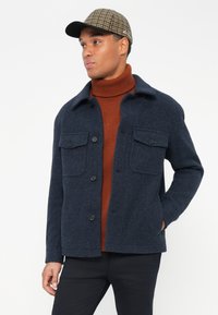 Muž v kárované čiapke, námorníckej bundovej sako s vreckami na klapke, hrdzavom rebrovanom roláku a čiernych nohaviciach, stojace proti bielej pozadie.