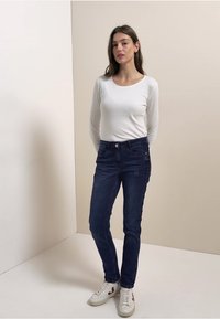 Valkoinen pitkähihainen paita, tummansiniset farkut ja valkoiset lenkkarit, joissa on punaisia yksityiskohtia. Farkut ovat kapeat ja niissä on hienovarainen pystysuora ommel.