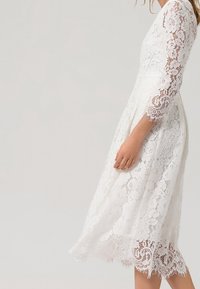 Bílé krajkové šaty s tříčtvrtečními rukávy, délka po kolena, květinový vzor a vroubkované okraje. Hladká vnitřní podšívka.