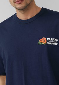 Navy-blaues Baumwoll-T-Shirt mit Rundhalsausschnitt, mit einem bunten Papaya-Graphic und dem Text "PAPAYA-flavored SURFWAX" auf der linken Brust.