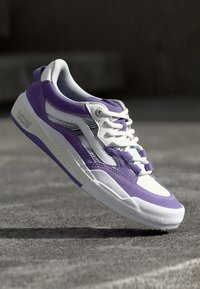 Zapatilla morada y blanca con parte superior de malla y ante, que presenta un diseño de franjas distintivo y cordones blancos, sobre un fondo gris.