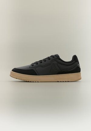 Czarny niski sneaker z beżową podeszwą, sznurowanym zapięciem i subtelnym tłoczonym logo, pokazany z boku na neutralnym tle.