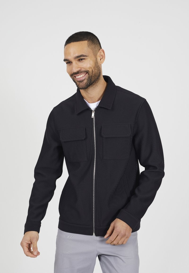 Schwarze, gerippte Zip-Jacke mit zwei Fronttaschen, Stehkragen und langen Ärmeln, getragen über einem weißen Hemd, vor einem schlichten Hintergrund gezeigt.