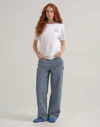Donna con lunghi capelli rossi che indossa una t-shirt bianca Lee, pantaloni larghi a righe blu e bianche e ciabatte blu, in piedi con le mani in tasca.